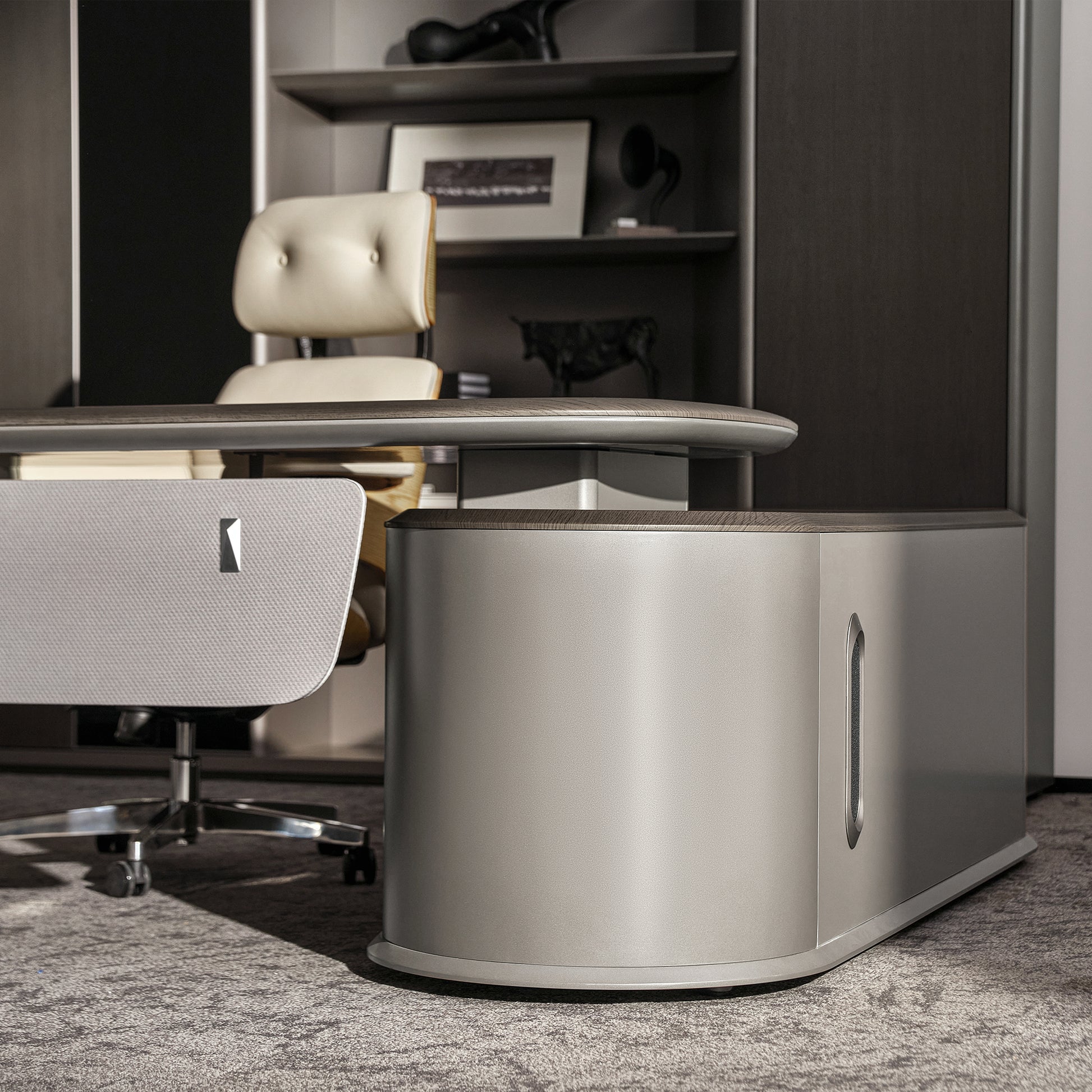 Eureka Ergonomic Zen Orion executive desk with wood grain top, matte gray curved base, cream leather chair, and bookcase in modern office.