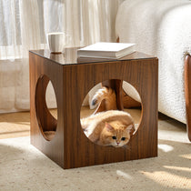 Eureka Ergonomic Wooden cat bed and side table with glass top, featuring a ginger cat peeking out from the opening and a book and mug on top.