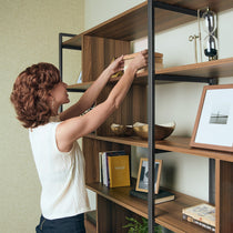 Woman Arranging Books On Napa Wood Bookcase Cabinet With Adjustable Shelves, Featuring Walnut Finish and Black Metal Frame.
