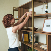 Woman Arranging Books On Napa Wood Bookcase Cabinet With Walnut Shelves, Brass Frame, and Decor.